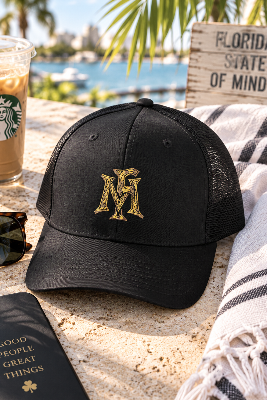 A black trucker-style baseball cap with a gold embroidered 'M' logo on the front, styled on a stone patio with a Starbucks iced coffee, sunglasses, a phone case, and a striped towel nearby.