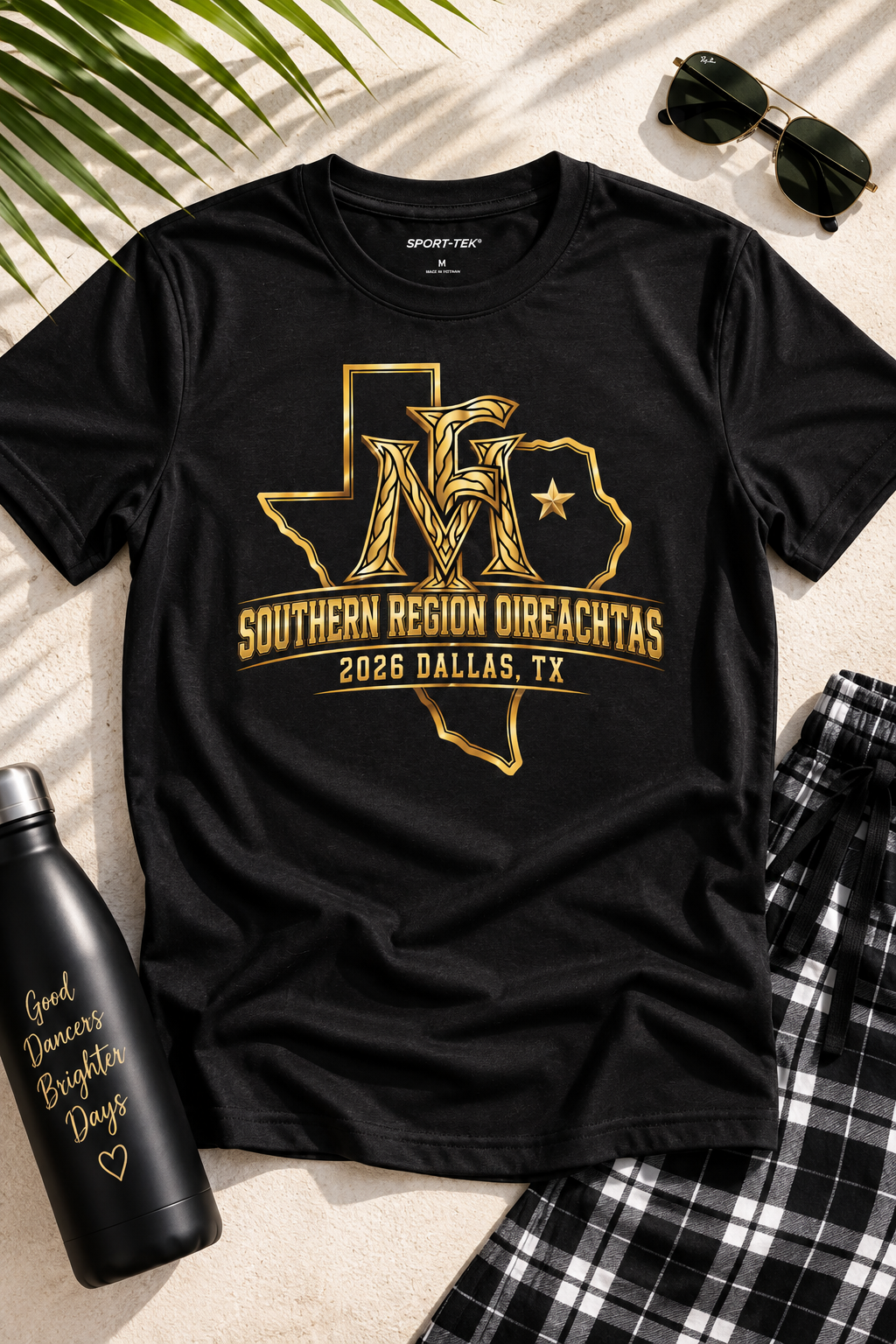 A black short-sleeve graphic T-shirt laid flat on a light beige surface with palm leaves, sunglasses, a black water bottle, and plaid shorts nearby. The shirt features a gold graphic of Texas with the letters 'M' and 'f' inside, plus the text 'SOUTHERN REGION DIREACHTAS' and '2026 DALLAS, TX'.