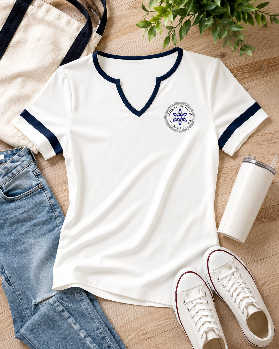 A white short-sleeve V-neck t-shirt with navy blue trim on the collar and sleeve cuffs, featuring a circular blue and gray emblem on the left chest. The shirt is styled in a flat lay on a light wood floor alongside blue jeans, a canvas tote bag, a white tumbler, white sneakers, and green leaves.