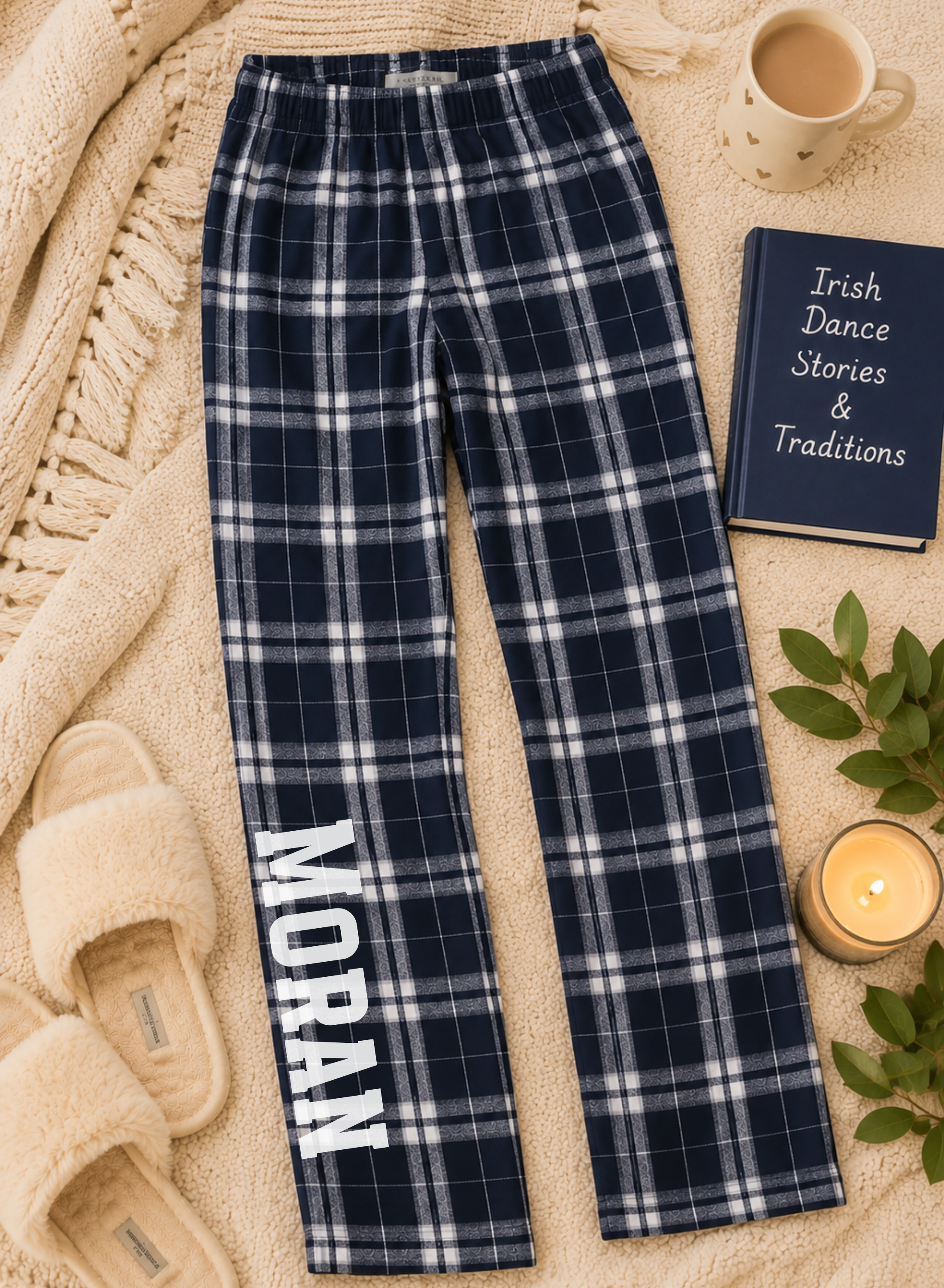 A pair of navy blue plaid pajama pants with the name 'MORAN' printed vertically in white on one leg, styled in a cozy flat-lay with a mug, book, candle, and slippers on a cream blanket.