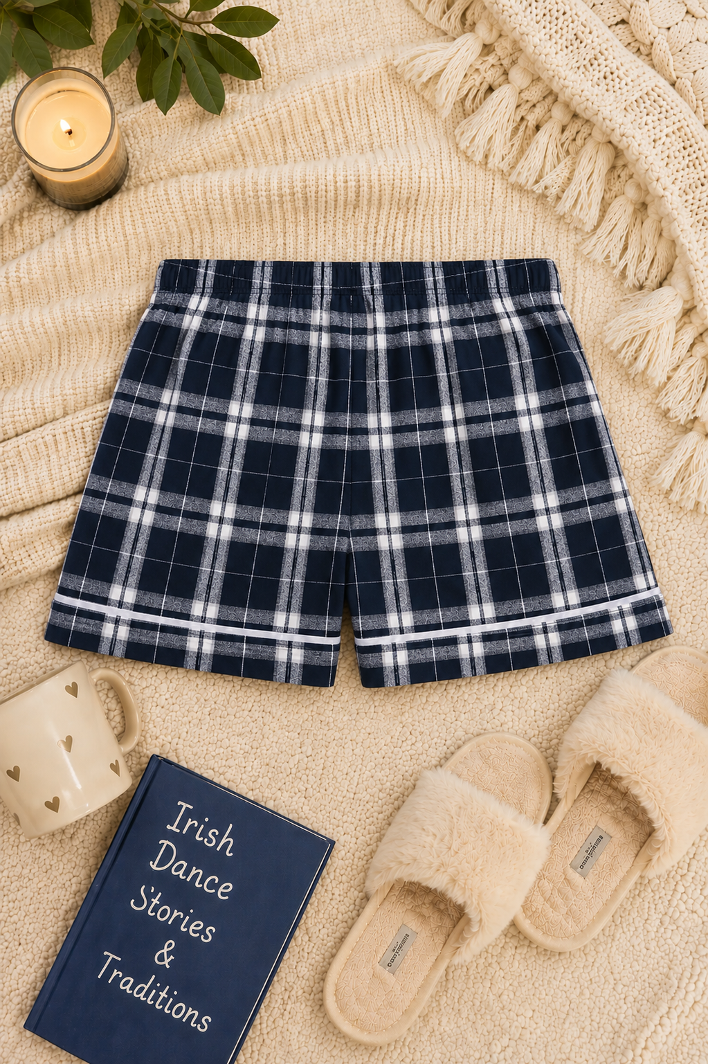 A pair of navy blue and white plaid shorts laid flat on a beige knit blanket, surrounded by cozy props including a candle, mug, book, slippers, and leaves.