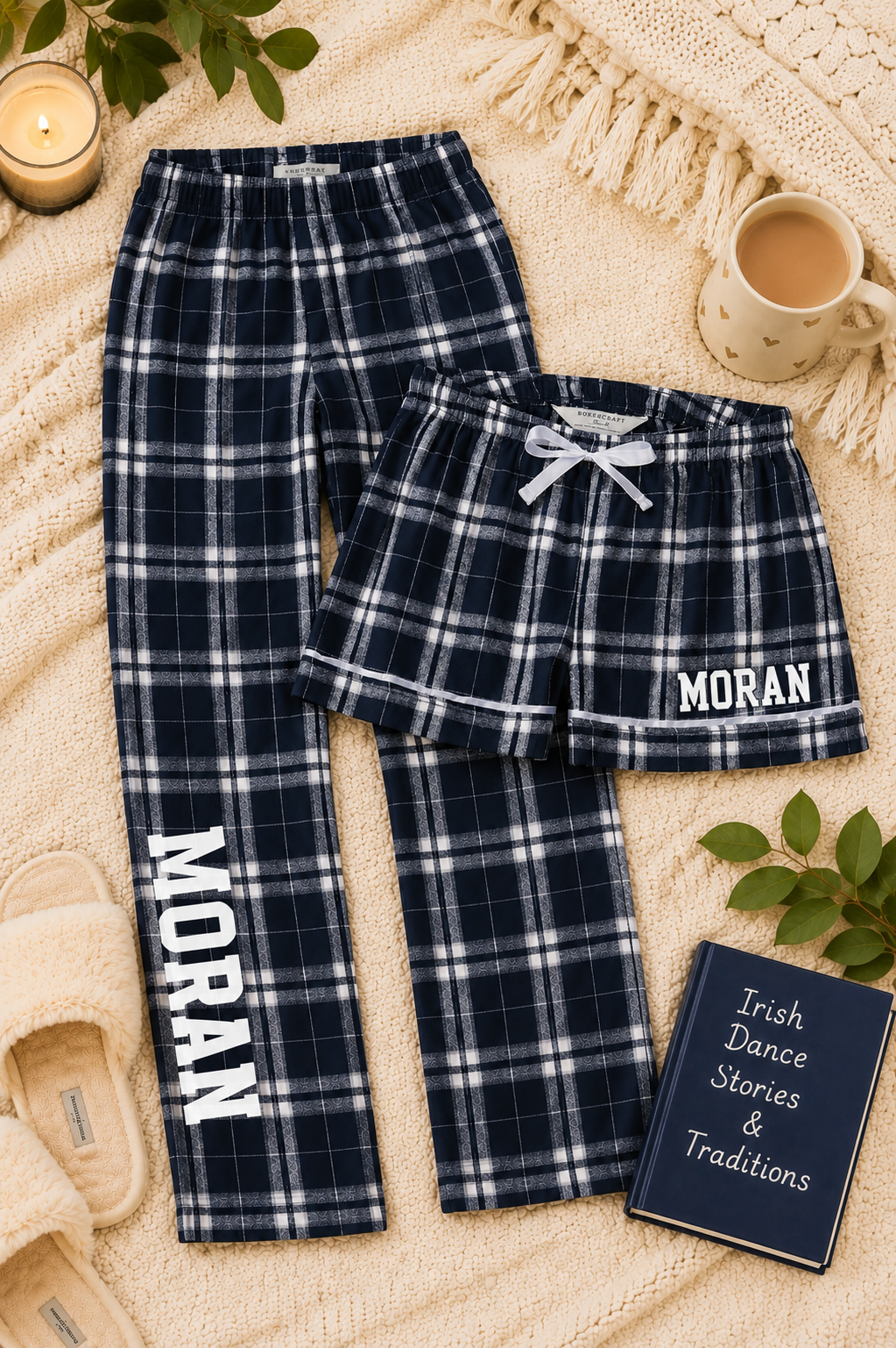 A matching pajama set laid flat on a cream knit blanket, including navy plaid pants with white vertical 'MORAN' text and navy plaid shorts with a white drawstring and 'MORAN' text on the leg. The scene includes a mug, candle, book, and small plant leaves as styled props.