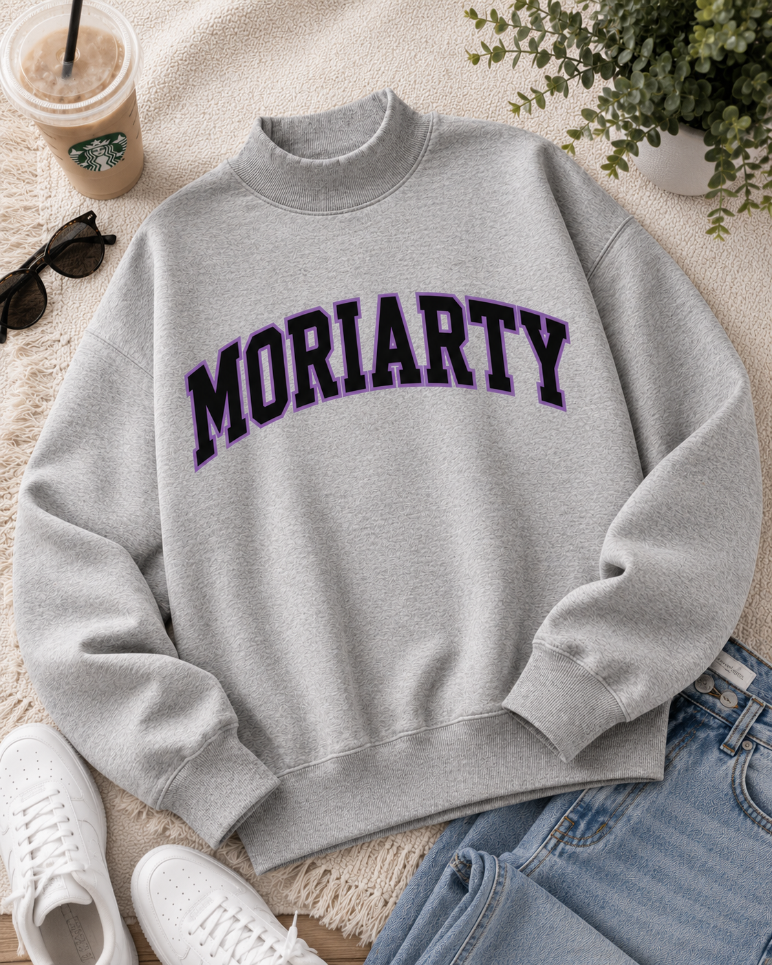 A light gray crewneck sweatshirt with 'MORIARTY' printed across the chest in dark purple varsity lettering, styled in a flat lay with jeans, white sneakers, sunglasses, an iced coffee, and a potted plant.