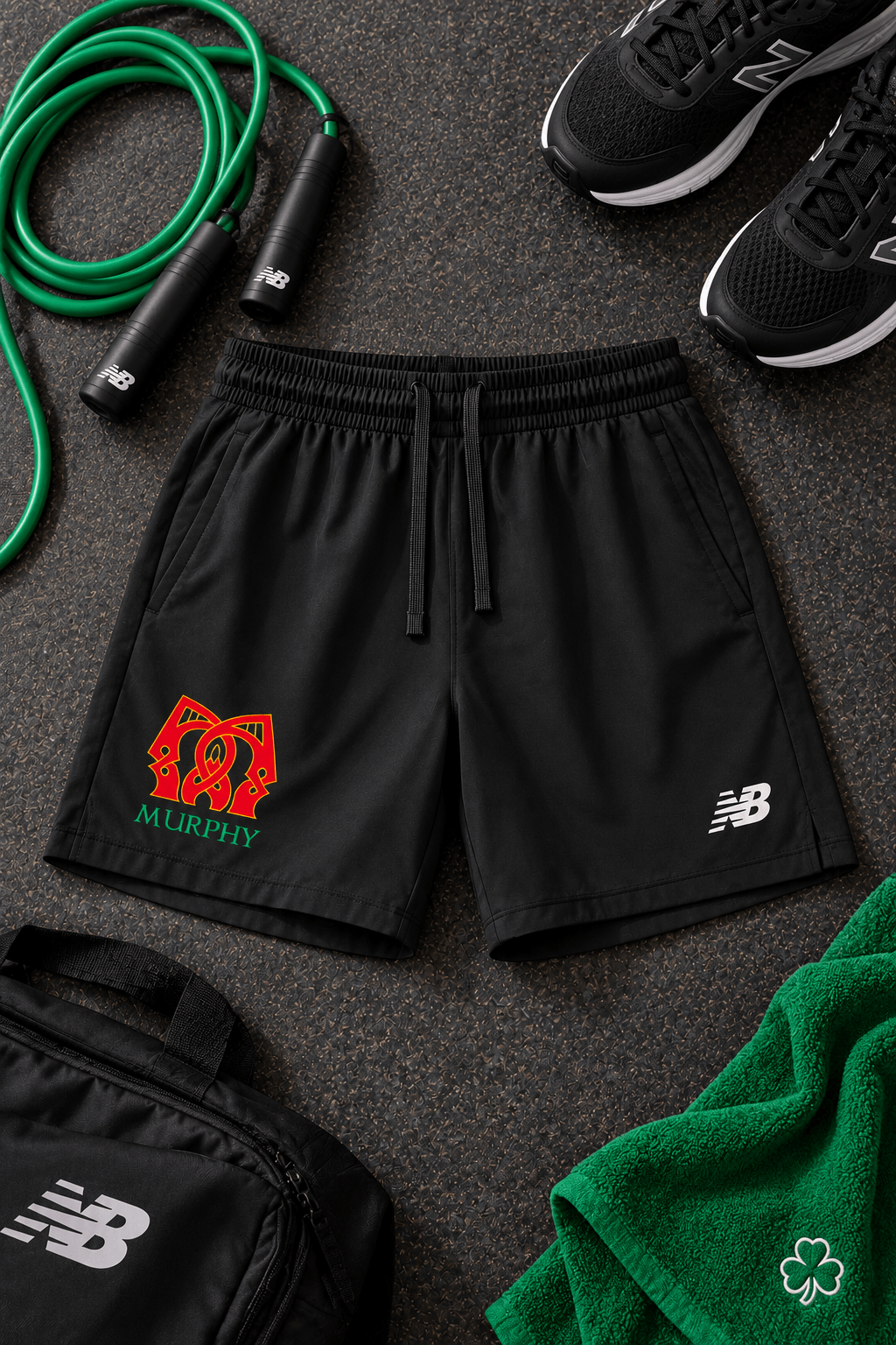 Black athletic shorts with a drawstring waist, featuring a red and green 'Murphy'  logo on the left and a white New Balance logo on the right, styled in a flat lay with a jump rope, shoes, a bag, and a towel.