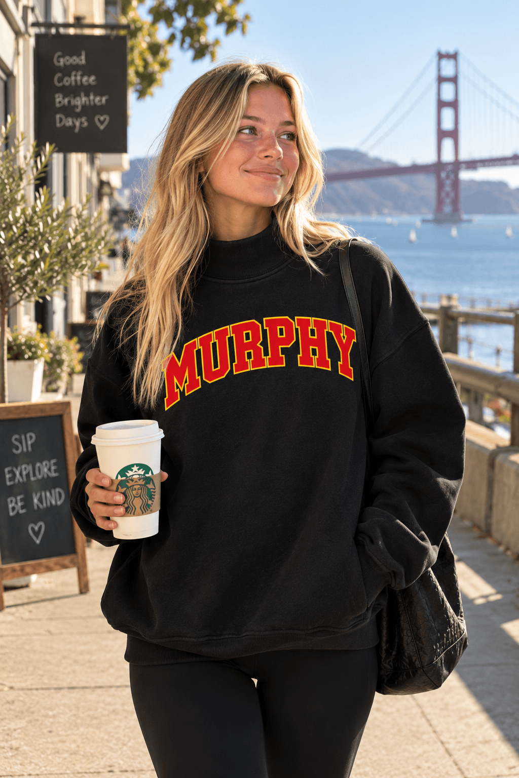 A woman stands on a sunny sidewalk in front of the Golden Gate Bridge, wearing a black sweatshirt with 'MURPHY' printed across the chest in red and yellow varsity-style lettering. She holds a Starbucks coffee cup.