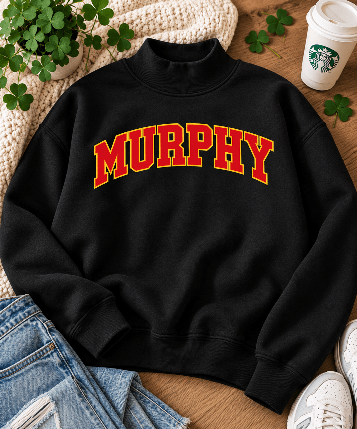 A black mock-neck sweatshirt with 'MURPHY' printed across the chest in red varsity-style letters with a yellow outline. The sweatshirt is styled in a flat-lay arrangement on a wooden surface with a knit blanket, jeans, a Starbucks cup, and shamrocks.