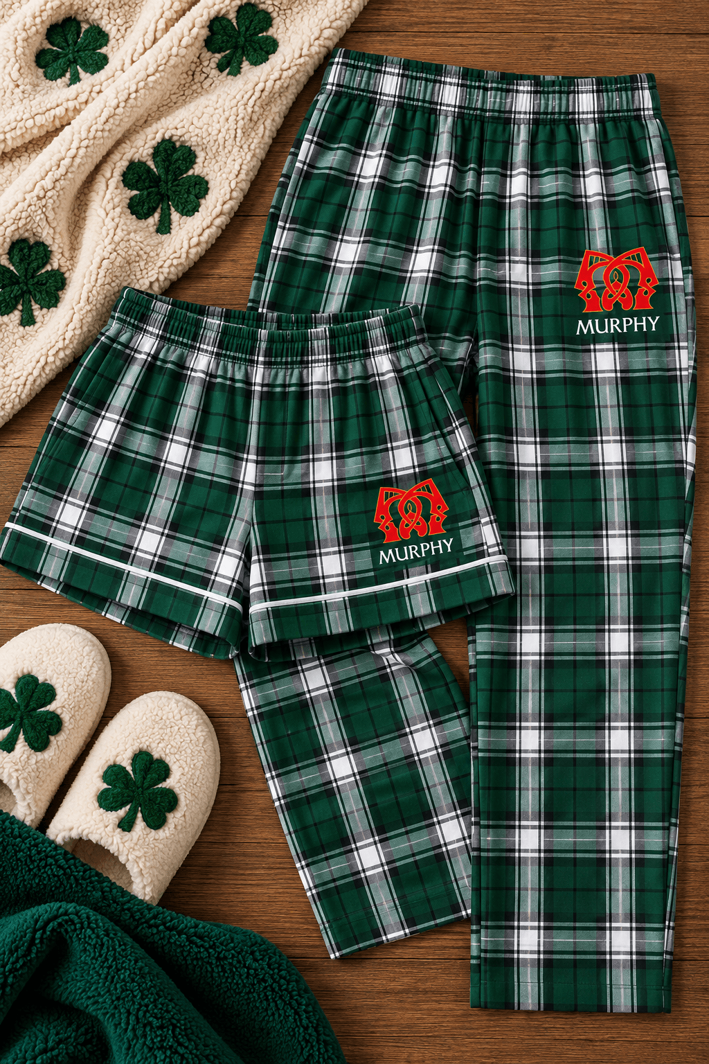 A matching pajama set laid flat on a wooden surface, including green plaid pajama pants and shorts with a red embroidered lion logo and the name 'MURPHY' on the left leg. The set is styled with a cream knit blanket, green shamrock accents, and matching slippers.