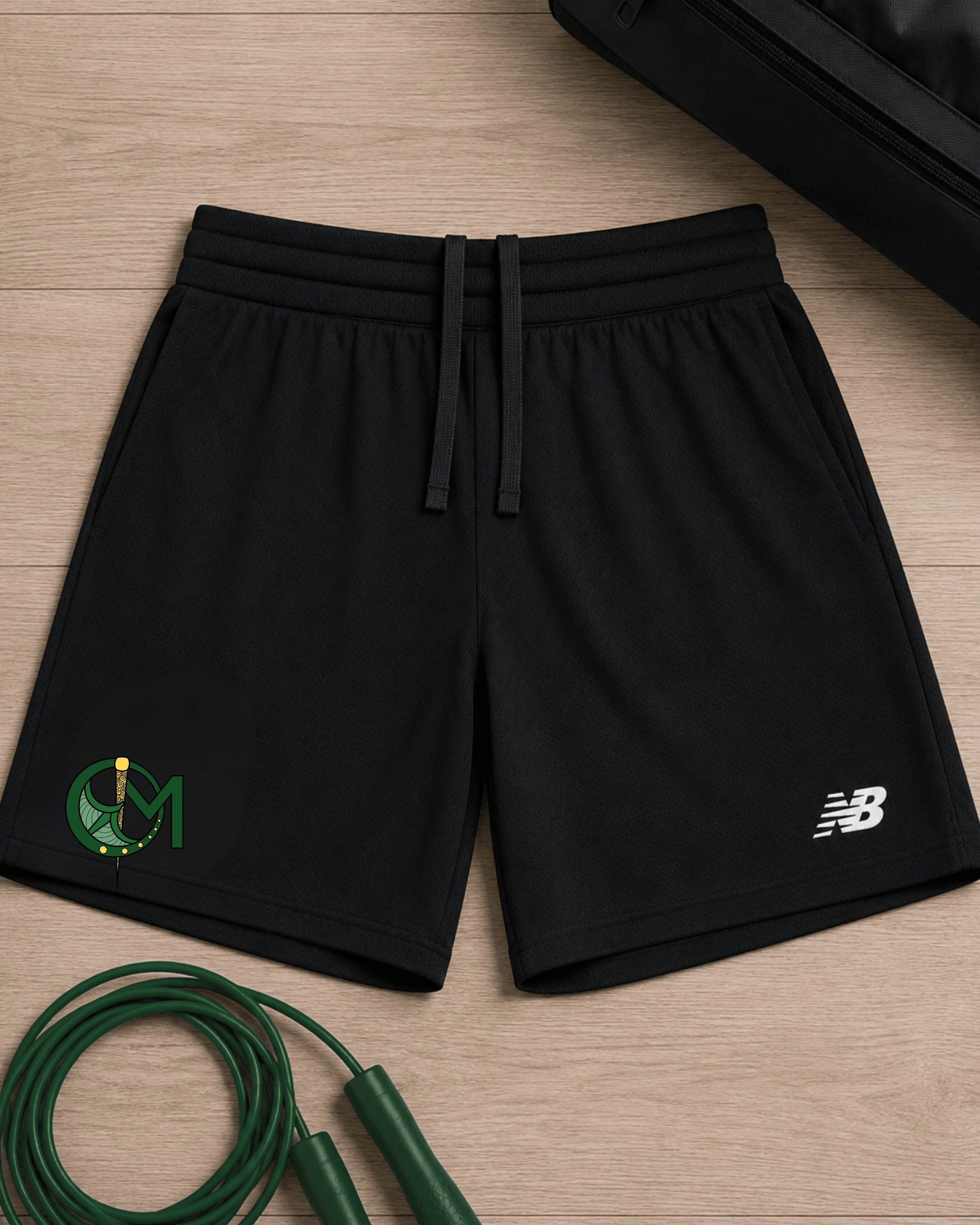 Black athletic shorts with a drawstring waist, featuring a green and yellow circular 'OM' logo on the left leg and a white New Balance 'NB' logo on the right leg, laid flat on a light wood background with a green jump rope and a black bag as props.
