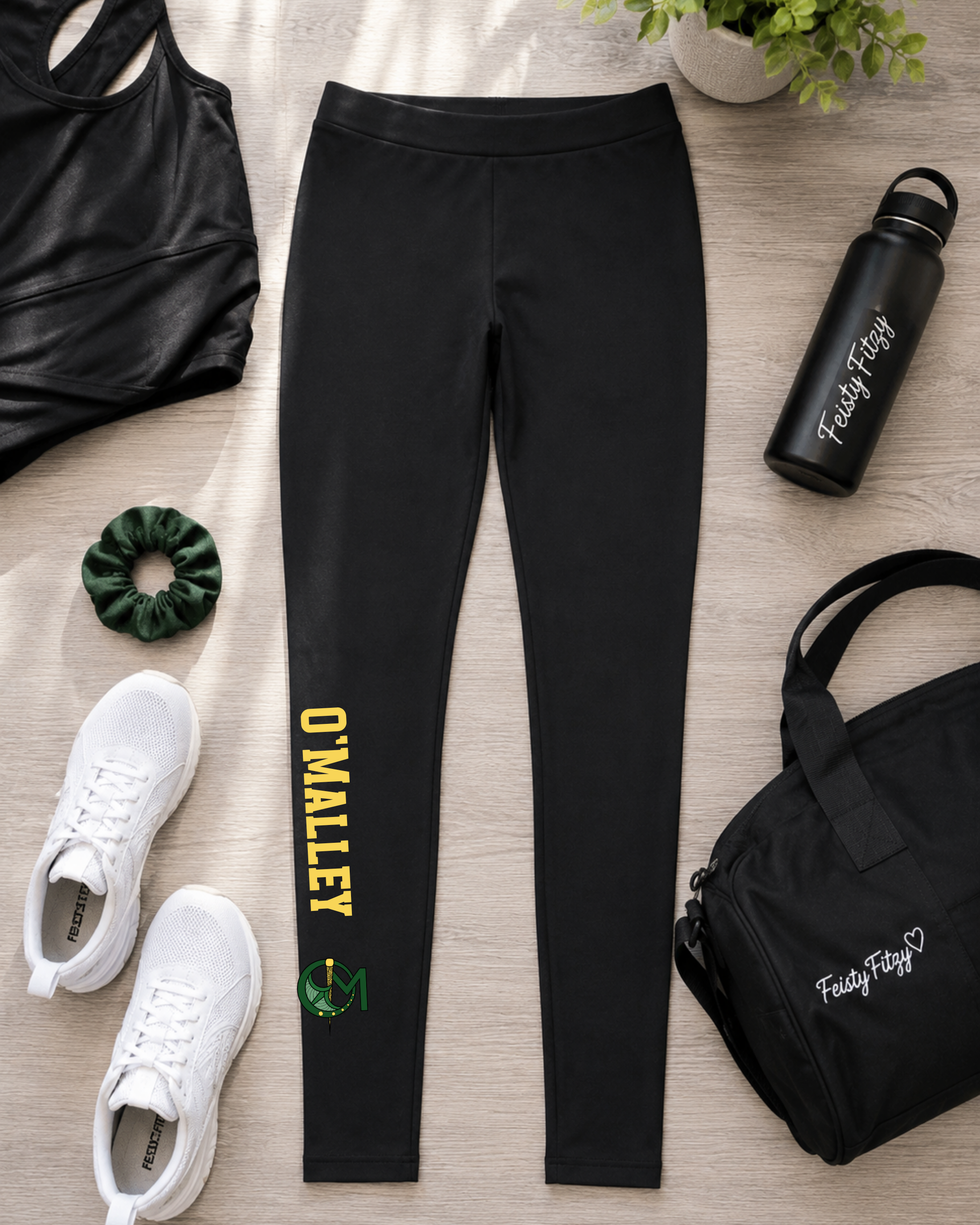 Black athletic leggings laid flat on a light wood surface, styled with a black sports bra, white sneakers, a green scrunchie, a black water bottle, and a black tote bag. The leggings have yellow 'O'MALLEY' text and a small green-and-white crest near the lower left leg.