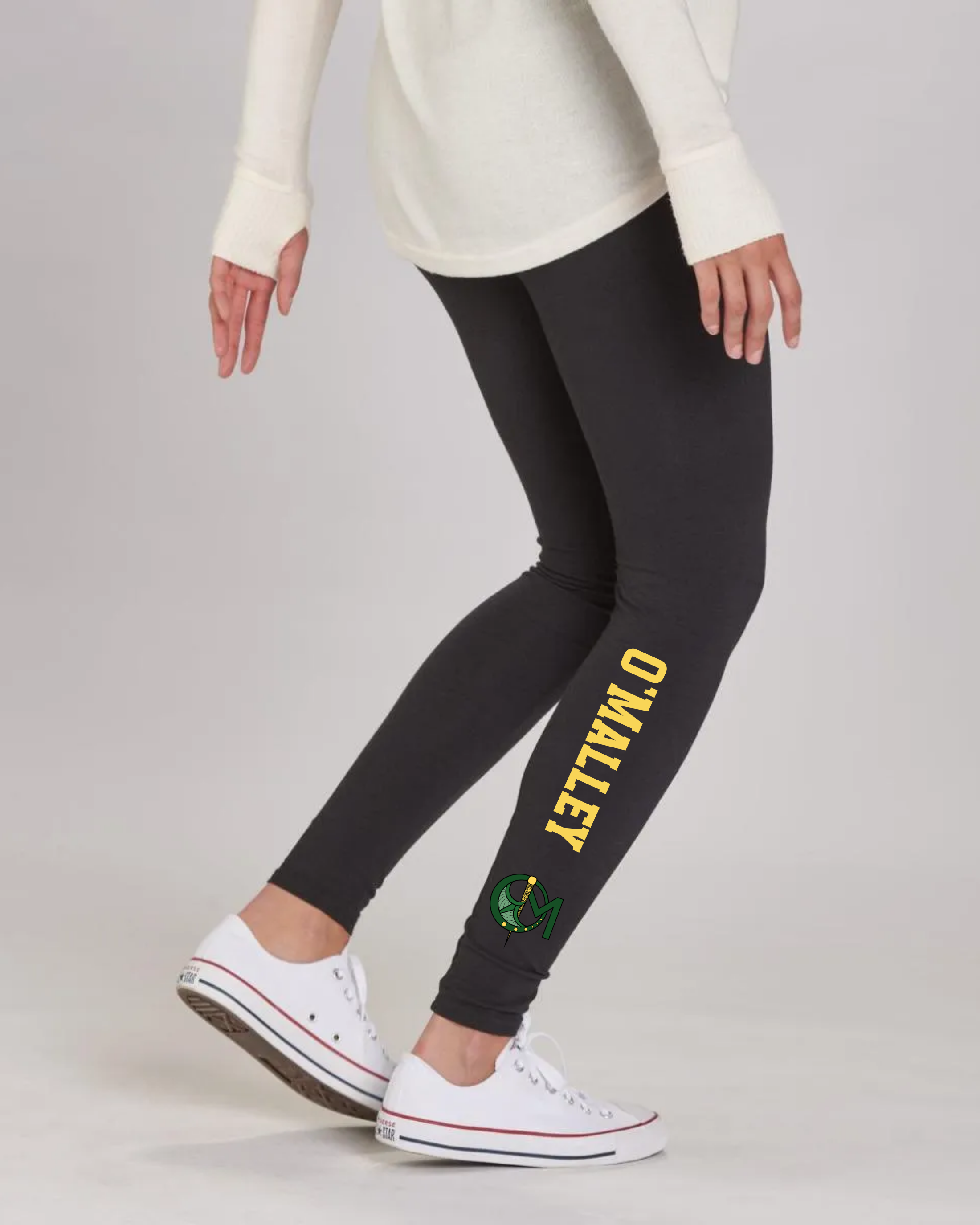 A person wearing black leggings with yellow 'O'MALLEY' text and a green logo on the lower leg, paired with white sneakers and a cream top, shown against a light gray studio background.