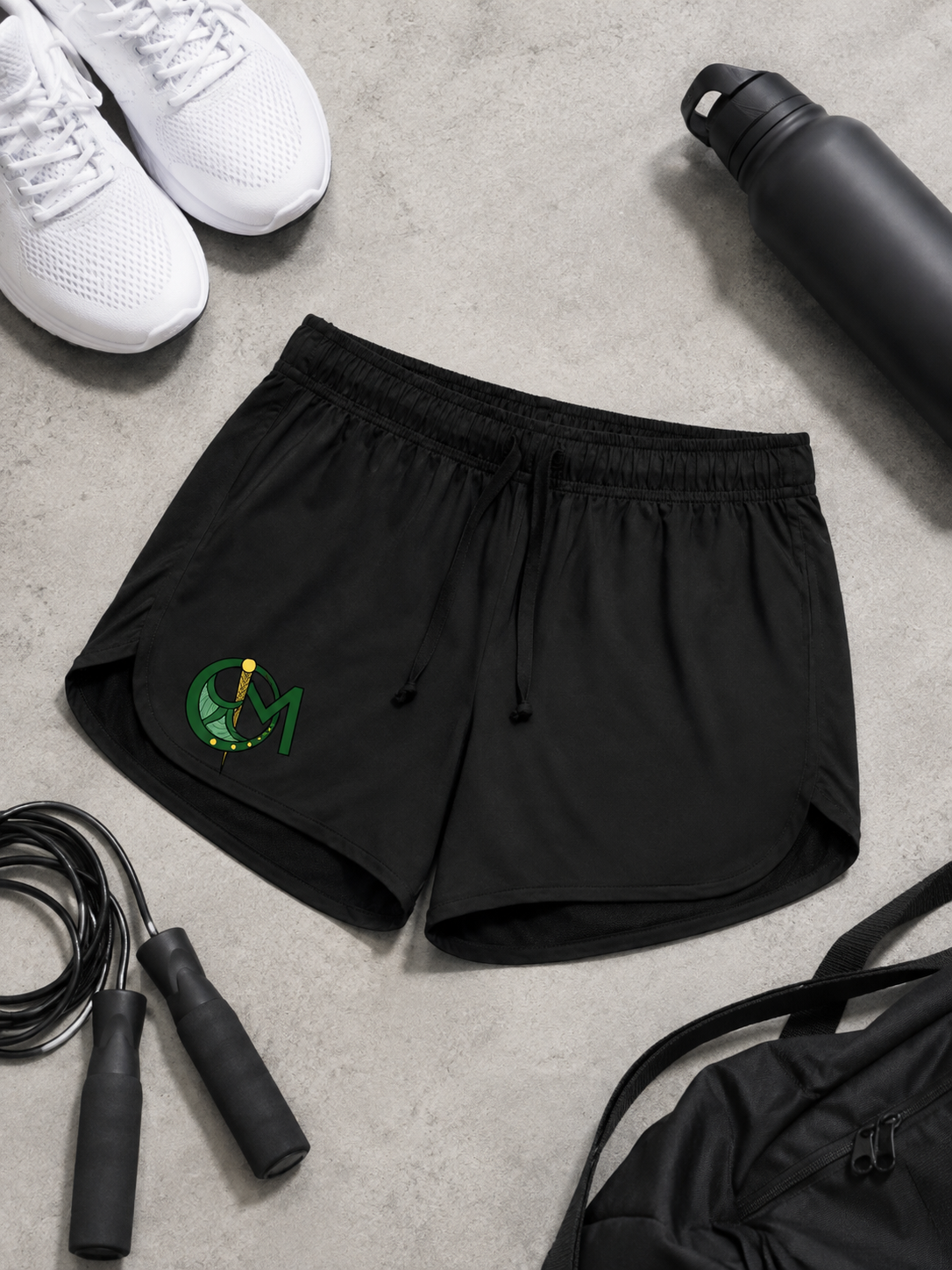 Black athletic shorts with a green and yellow circular logo on the left leg, laid flat on a gray concrete surface surrounded by white sneakers, a black water bottle, a jump rope, and a black backpack.
