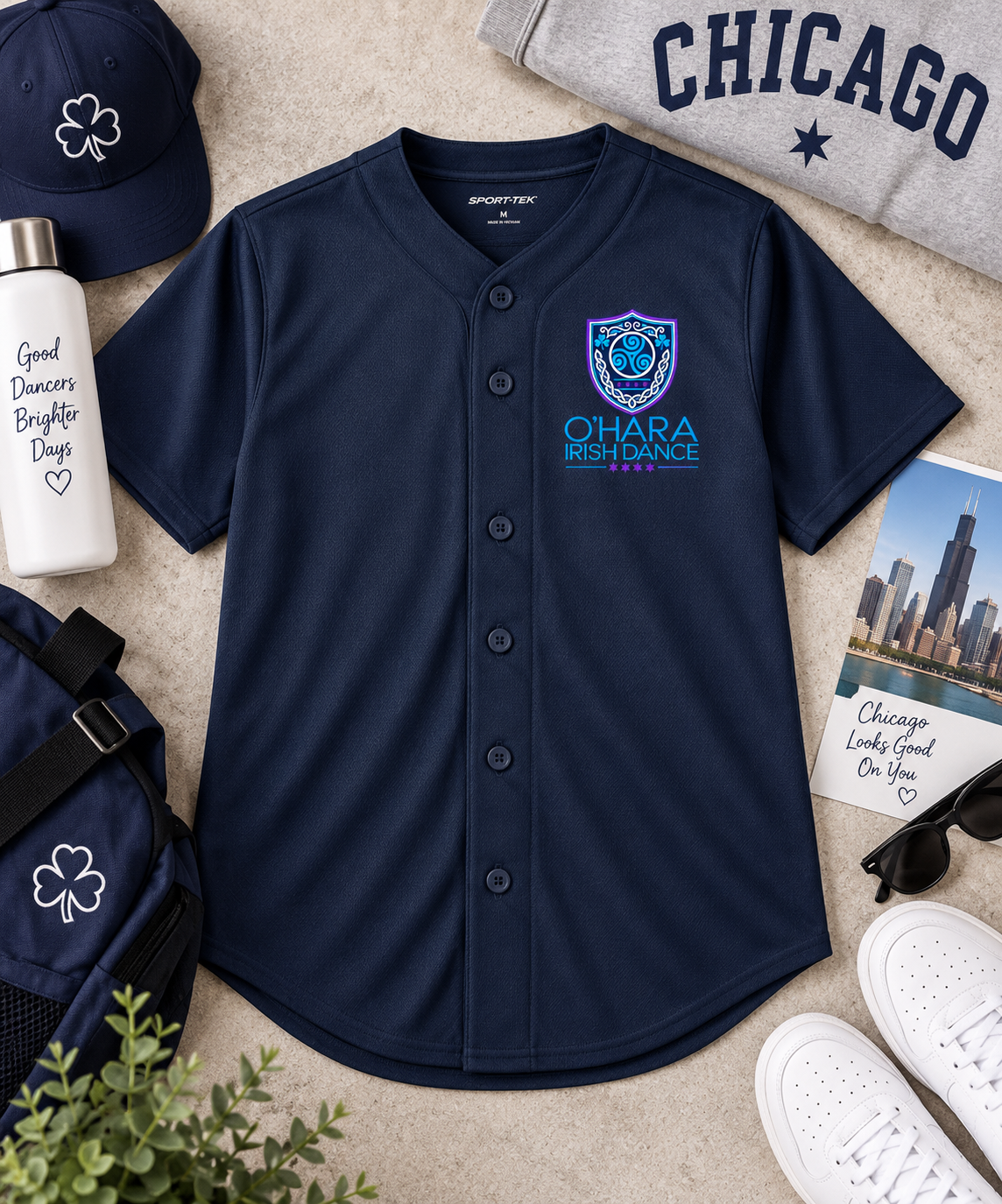 A navy blue baseball-style button-front jersey laid flat on a light beige surface, surrounded by styled props including a gray sweatshirt with 'CHICAGO', a white bottle, a navy cap, a navy backpack, sunglasses, white sneakers, and a small plant. The jersey has a shield crest on the left chest with 'OHARA IRELAND' text.
