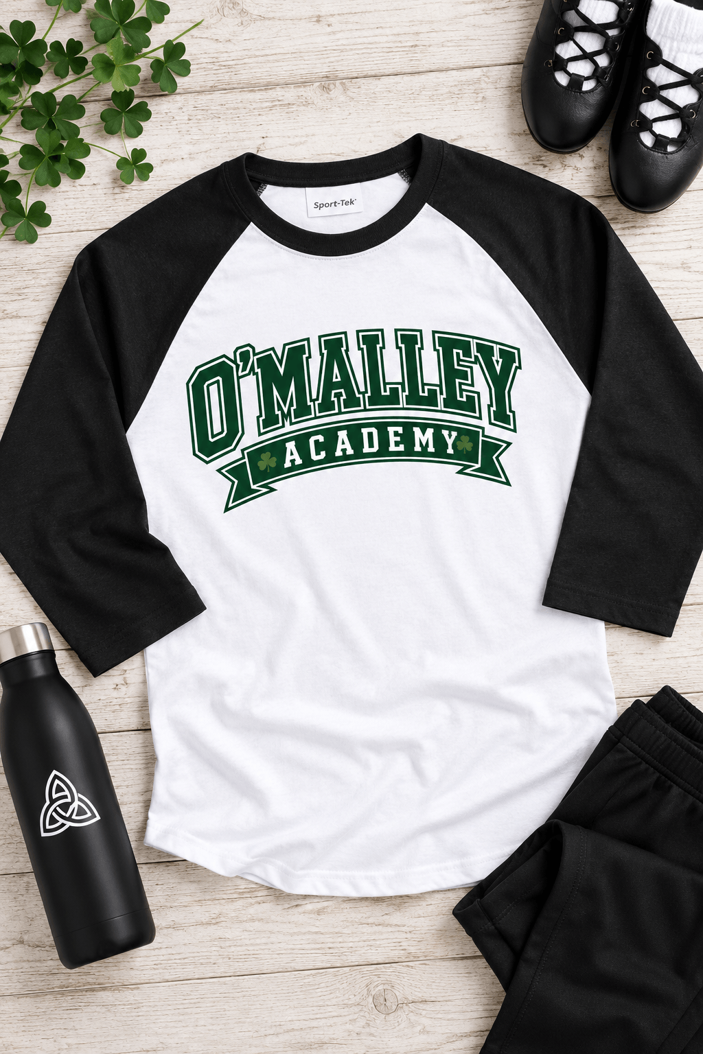 A white and black raglan baseball t-shirt with 'O'MALLEY ACADEMY' printed in green on the front, styled in a flat lay with black shorts, a black water bottle, black shoes, and green clover on a light wood background.