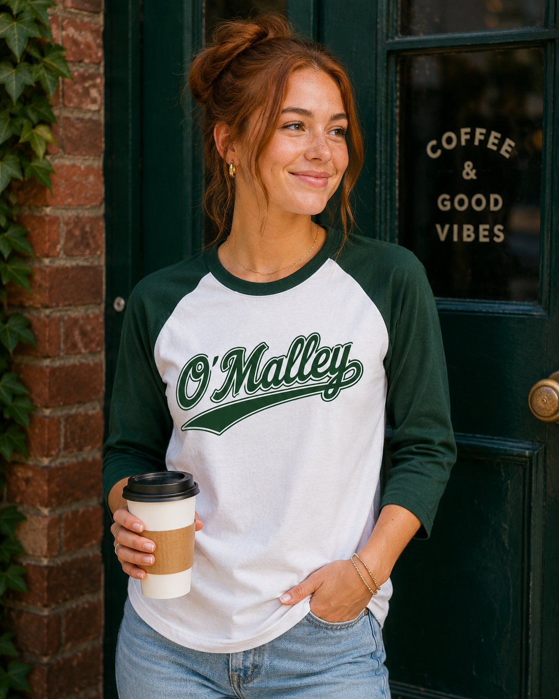 A woman wearing a white and dark green raglan baseball t-shirt with 'O'Malley' printed in green script across the chest, holding a coffee cup in front of a dark green cafe door.