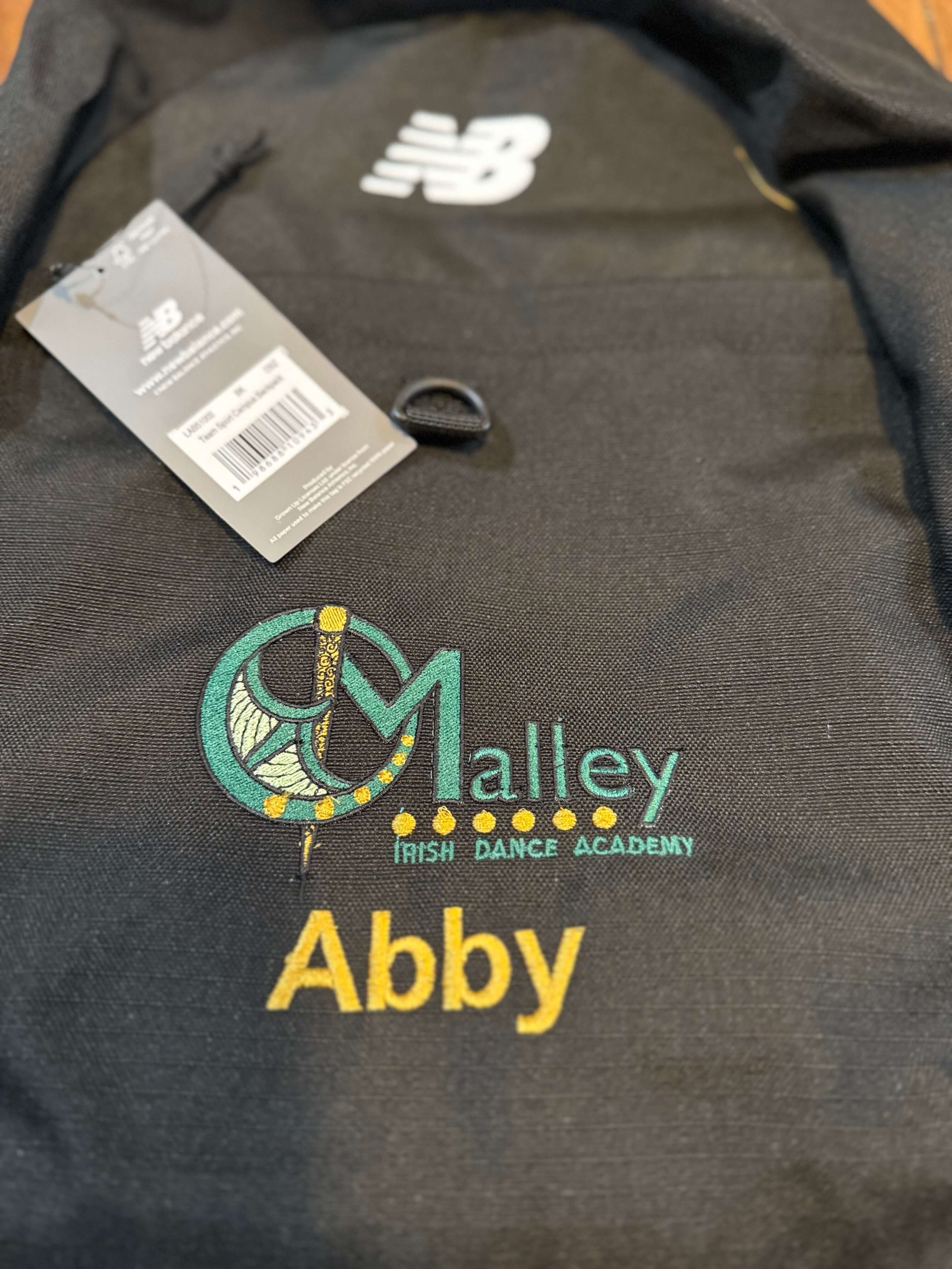A close-up of a dark gray New Balance pullover hoodie featuring custom embroidery. The embroidery includes a teal and yellow 'CMalley Irish Dance Academy' logo and the name 'Abby' in yellow.