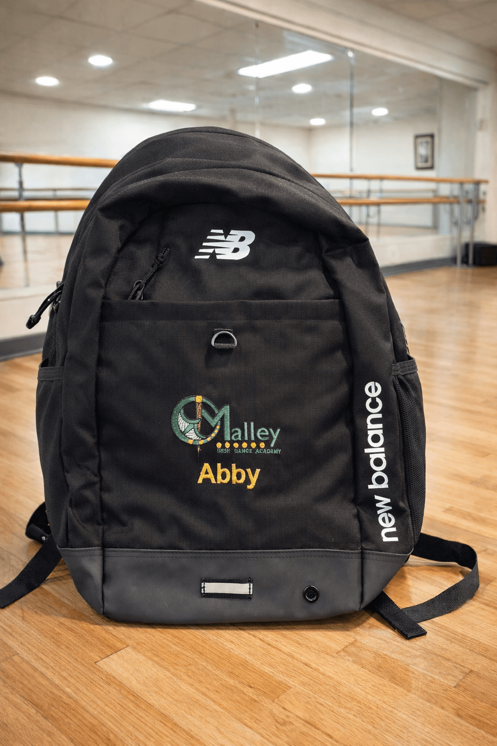 A black New Balance backpack with custom embroidery on a wooden floor inside a dance studio. The backpack features a white New Balance logo on the upper back, a front pocket with a green and gold Malley Skates logo and the name 'Abby' in yellow, and 'new balance' printed vertically in white on the side strap.