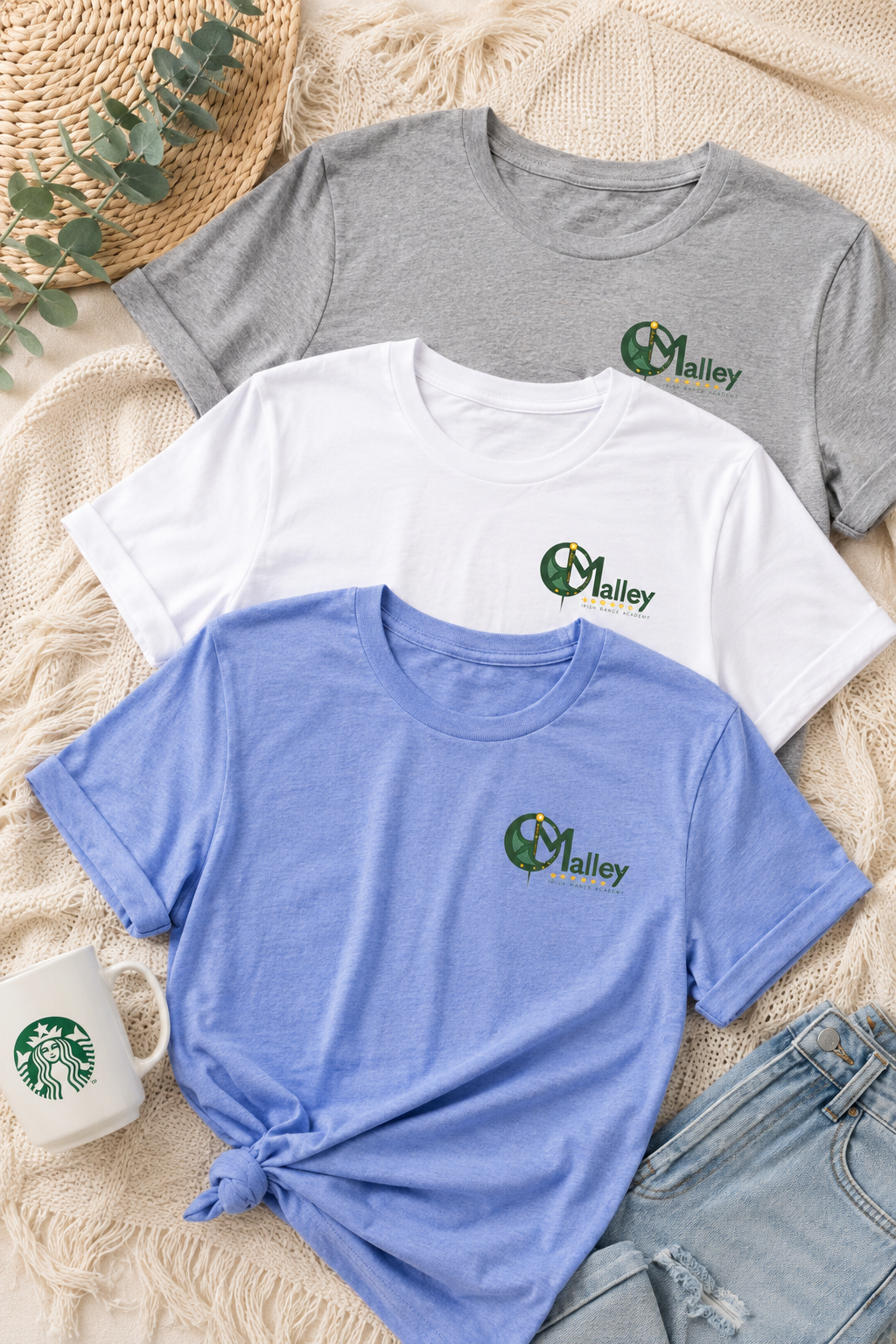 Three short-sleeve crewneck T-shirts in gray, white, and light blue, laid flat on a beige knit blanket with a woven hat, eucalyptus leaves, a white Starbucks mug, and denim shorts. Each shirt has a green and white 'O'Malley' logo printed on the left chest.