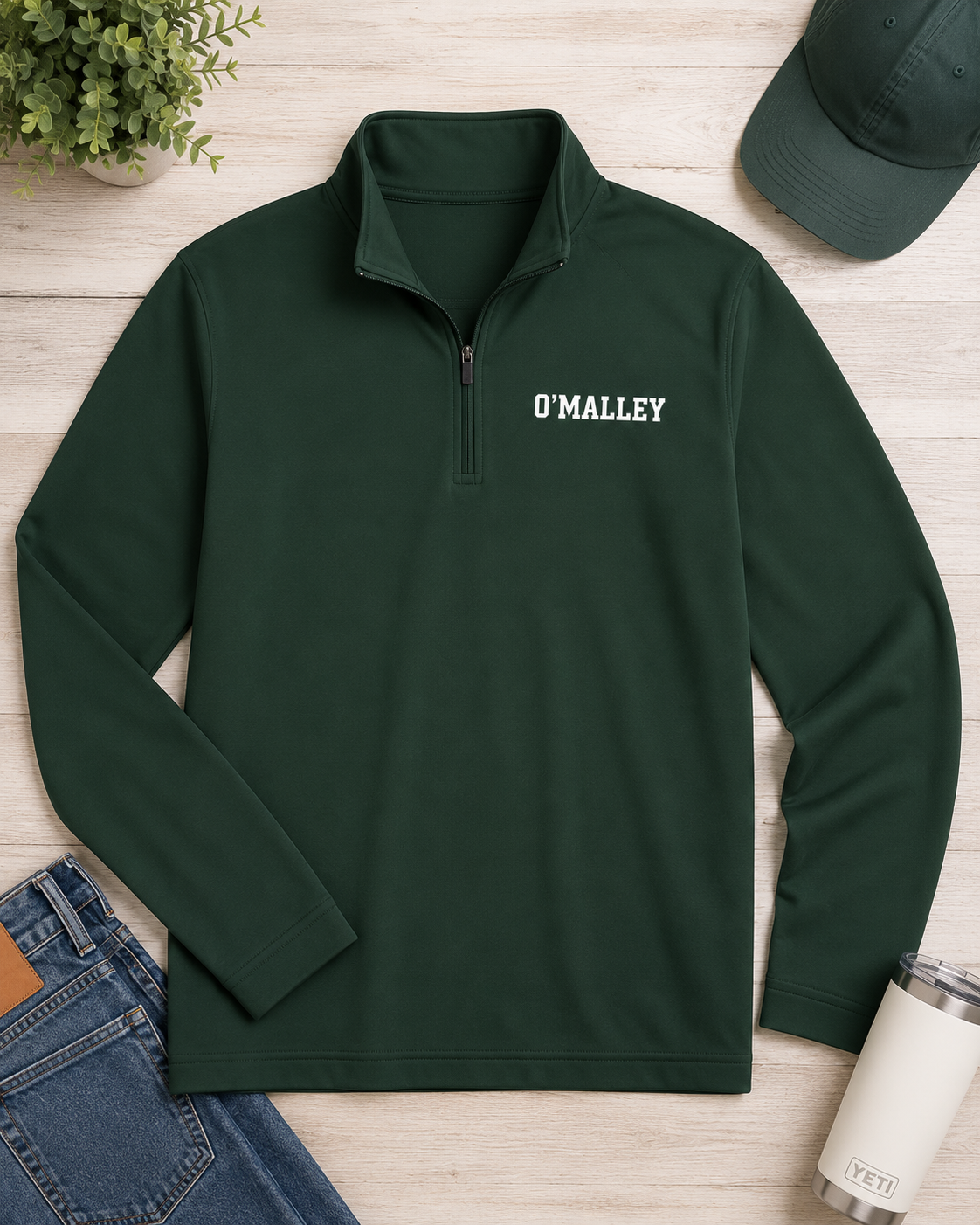 A dark green long-sleeve quarter-zip pullover with 'O'MALLEY' printed in white on the left chest, styled in a flat lay on a light wood background with jeans, a cap, a plant, and a tumbler.