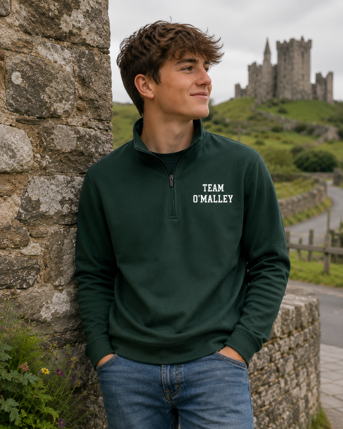 A young man wearing a dark green quarter-zip sweatshirt with 'TEAM O'MALLEY' printed in white on the chest, standing outdoors next to a stone wall with a castle in the background.