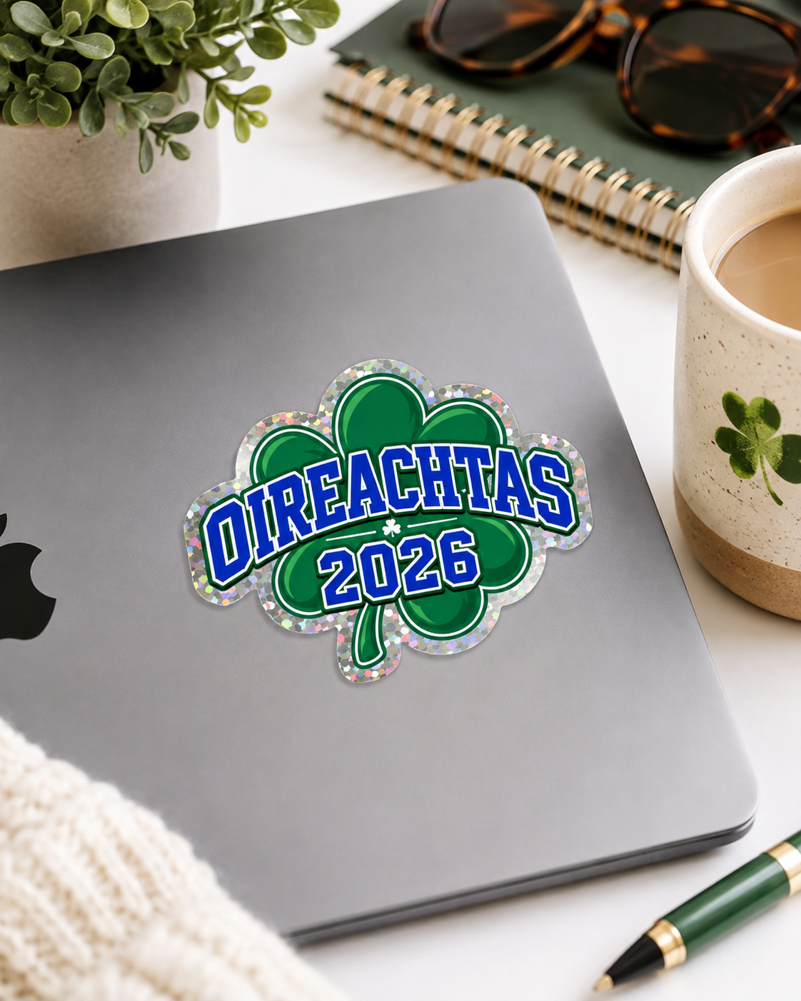 A St. Patrick's Day themed die-cut sticker featuring a green shamrock outline and the text 'OIREACHTAS 2026' in blue with a glittery silver border, shown applied to the back of a silver laptop on a desk with a plant, notebook, sunglasses, and a coffee mug.