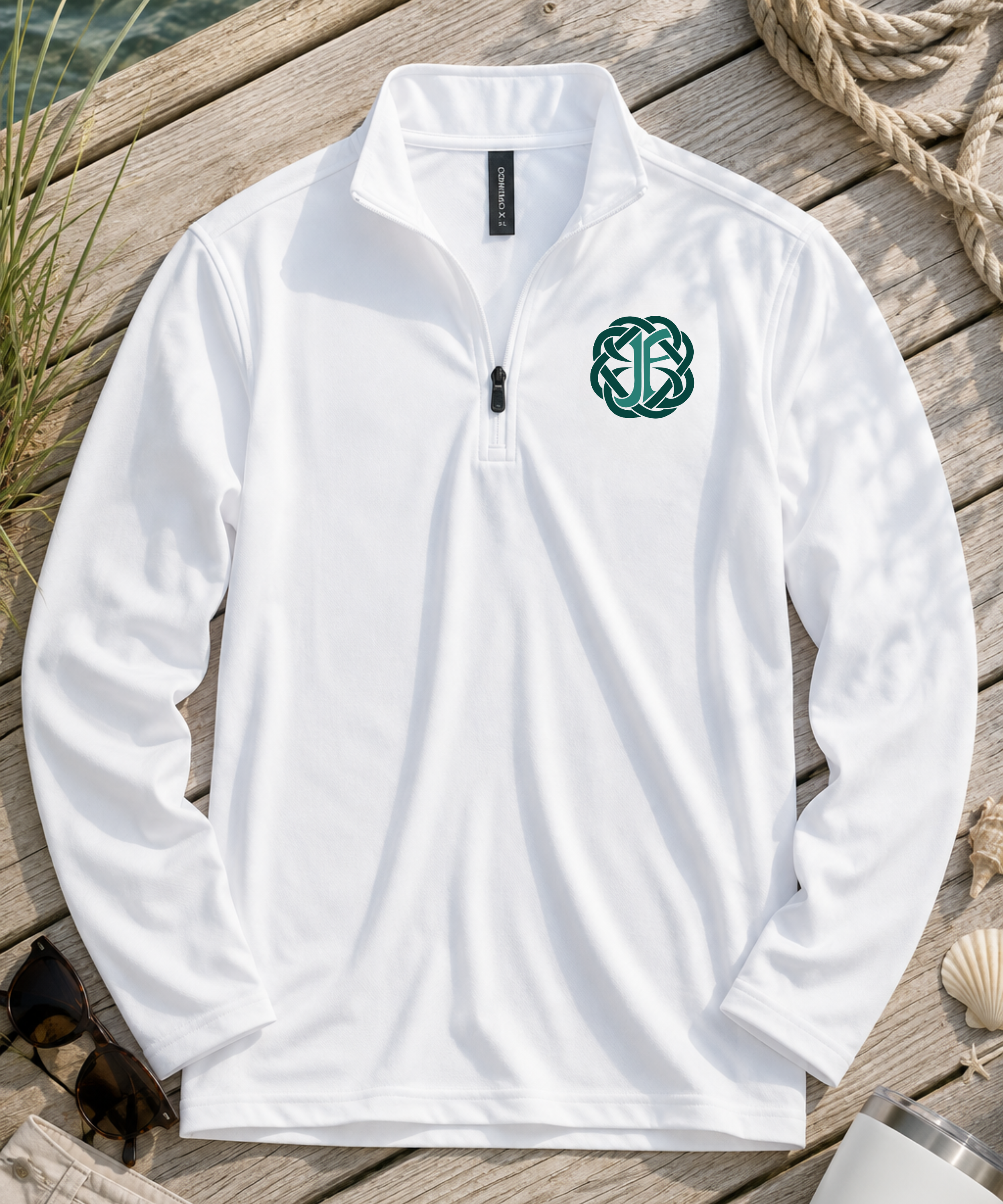 A white long-sleeve quarter-zip pullover laid flat on a wooden dock, featuring a green Celtic knot emblem on the left chest. The background includes natural bohemian props like rope, seashells, and seaweed.