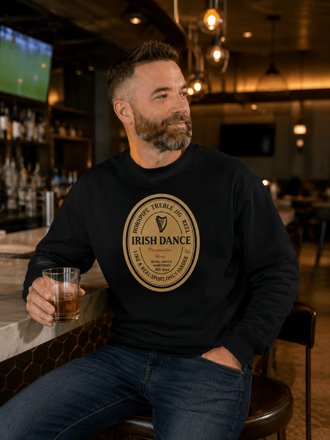 A man sitting at a bar wearing a black crewneck sweatshirt with a large circular gold and white 'Irish Dance' logo on the chest, holding a glass of amber beer.