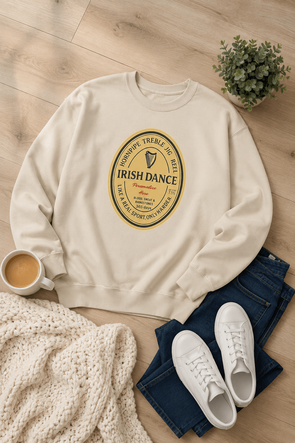 A cream-colored crewneck sweatshirt with a circular 'Irish Dance' graphic printed on the chest, styled in a flat lay on a light wood floor with blue jeans, white sneakers, a knit blanket, a coffee cup, and a small potted plant.