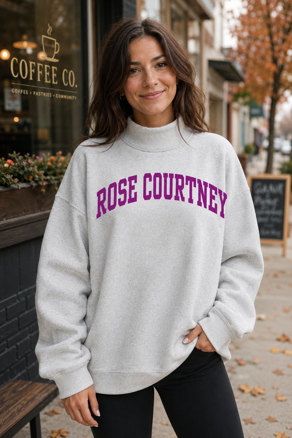 A woman wearing a light heather gray mock neck sweatshirt with 'ROSE COURTNEY' printed in purple arched letters across the chest, standing on a city sidewalk in front of a coffee shop.
