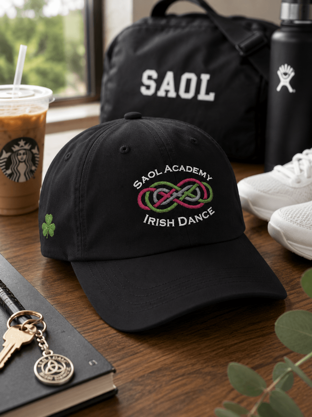 A black baseball cap with 'SAOL ACADEMY IRISH DANCE' embroidered in white and a green/red infinity symbol on the front, styled on a wooden table with a Starbucks iced coffee, a black duffel bag, white sneakers, and a keychain.