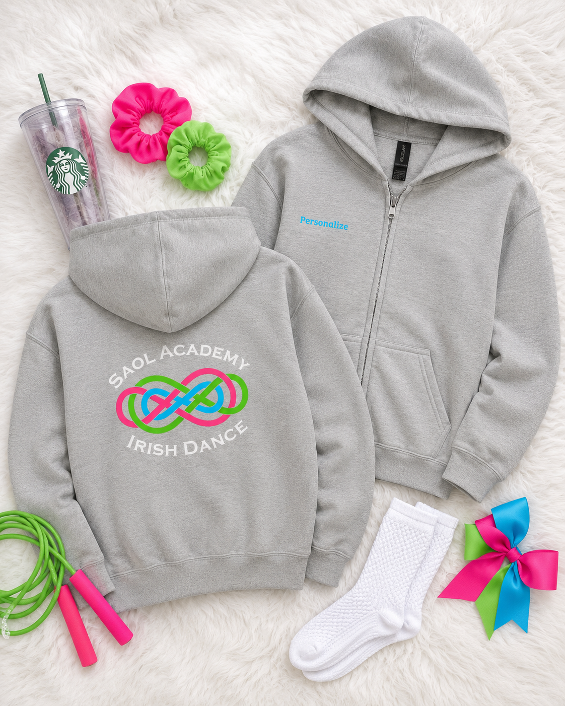 A flat lay of a light gray zip-up hoodie shown from both front and back, styled with a Starbucks cup, pink and green scrunchies, a jump rope, white socks, and a pink and blue bow on a white faux fur background. The back of the hoodie features a colorful graphic of interlocking dance rings with the text 'SAOL ACADEMY IRISH DANCE'.
