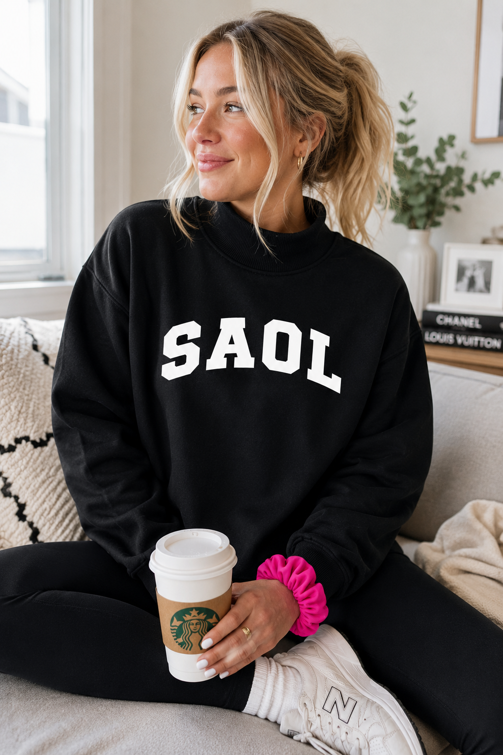A woman sitting on a couch wearing a black sweatshirt with 'SAOL' printed in white block letters across the chest. She is holding a Starbucks coffee cup and wearing white sneakers with a bright pink scrunchie on her wrist.