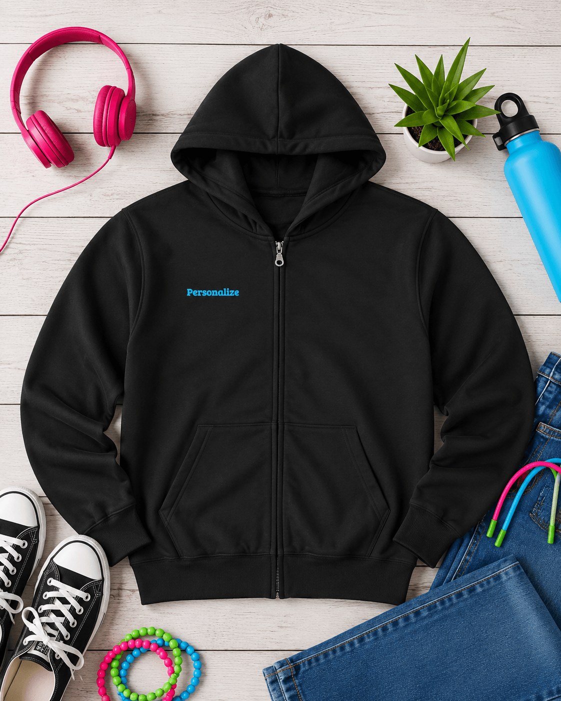 A black zip-up hoodie laid flat on a white wood background, surrounded by colorful lifestyle props including pink headphones, a small green plant, a blue water bottle, black sneakers, jeans, and bright bracelets. The hoodie has a small blue 'Personalize' text print on the left chest.