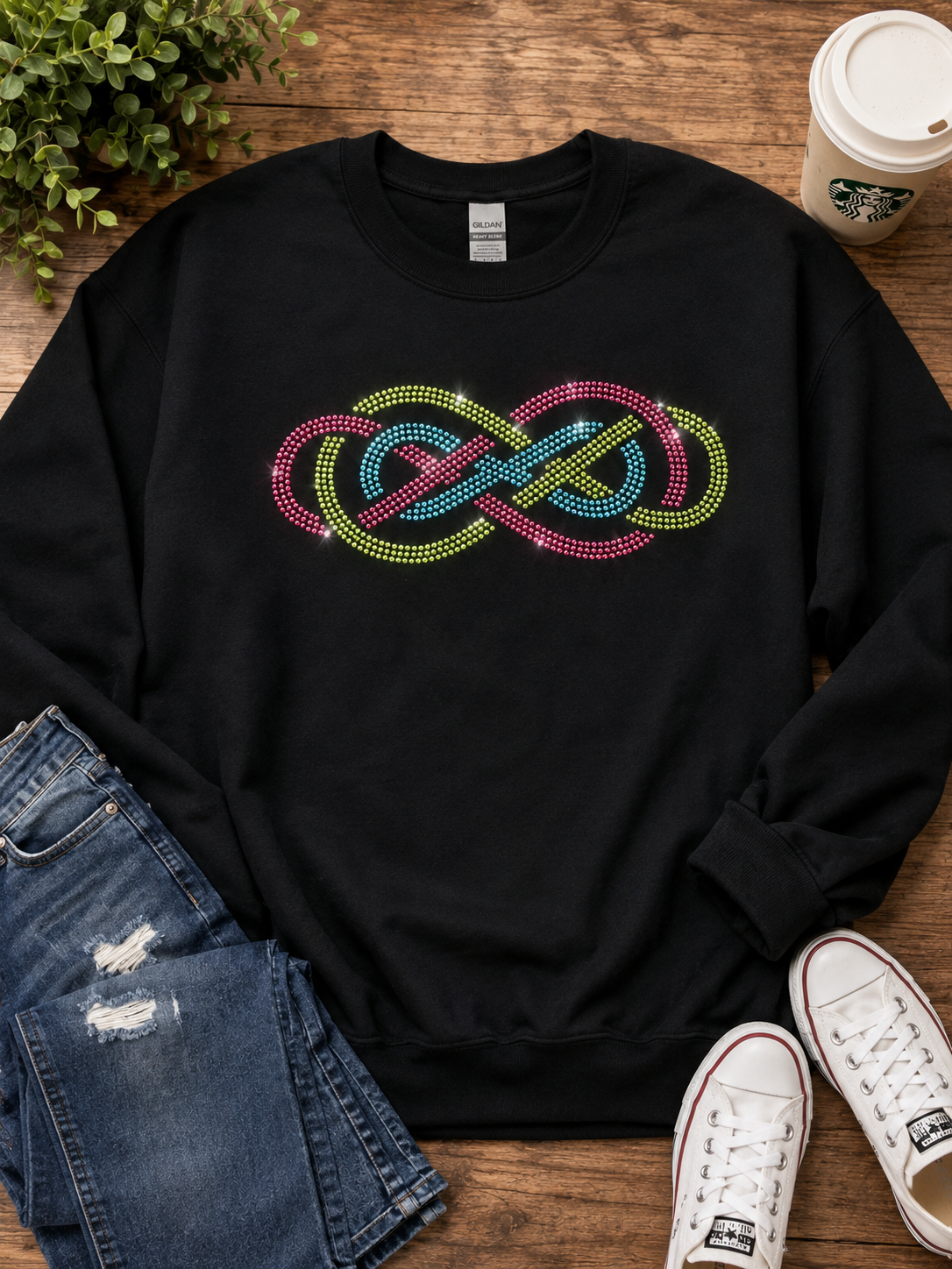A black crewneck sweatshirt with a colorful rhinestone infinity knot graphic on the chest, styled in a flat lay with blue jeans, white sneakers, a Starbucks coffee cup, and a green plant on a wooden surface.
