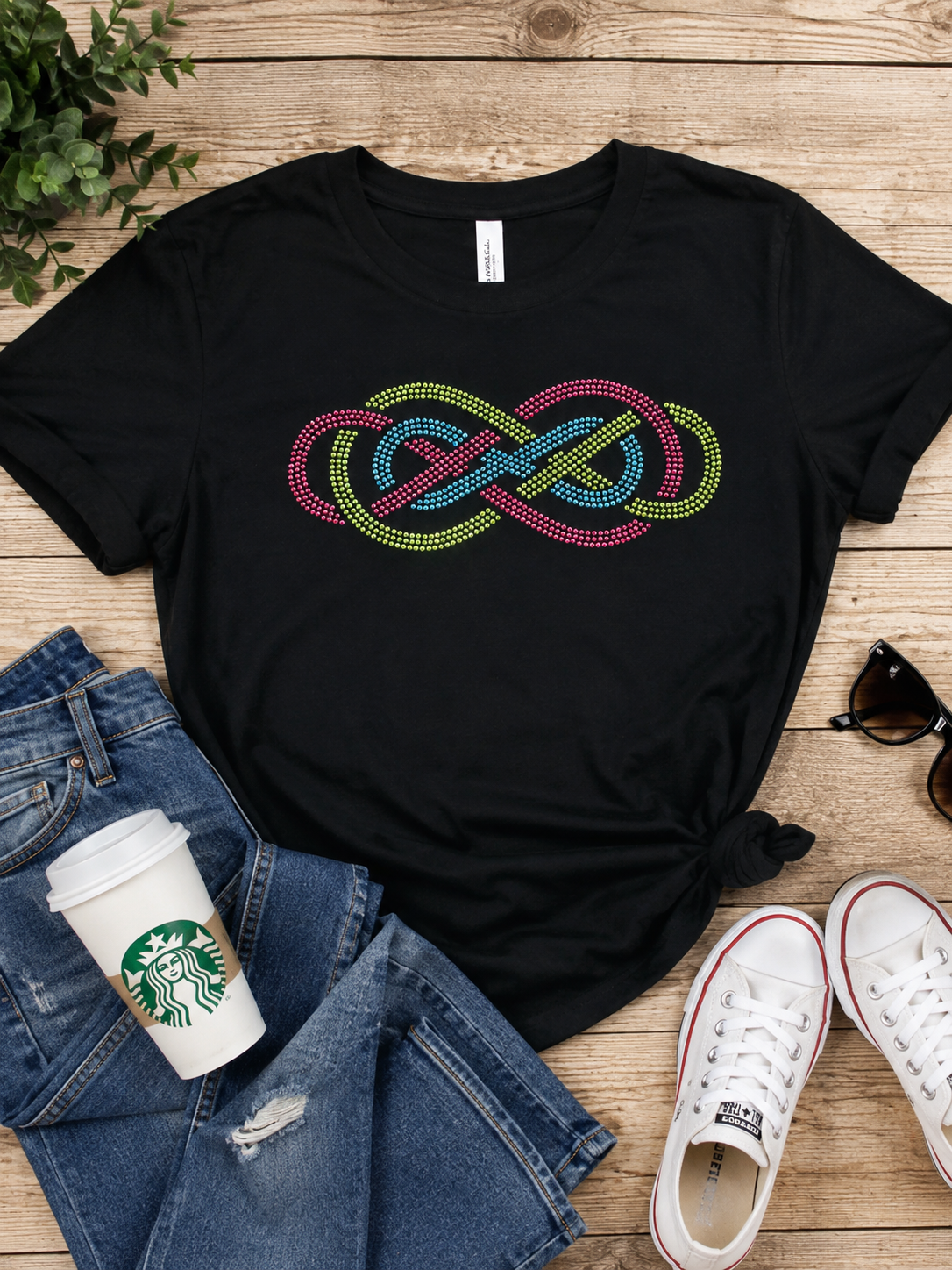 A black short-sleeve T-shirt laid flat on a wooden surface, featuring a colorful dotted infinity knot graphic across the chest. The shirt is styled with blue jeans, a Starbucks cup, white sneakers, and sunglasses.