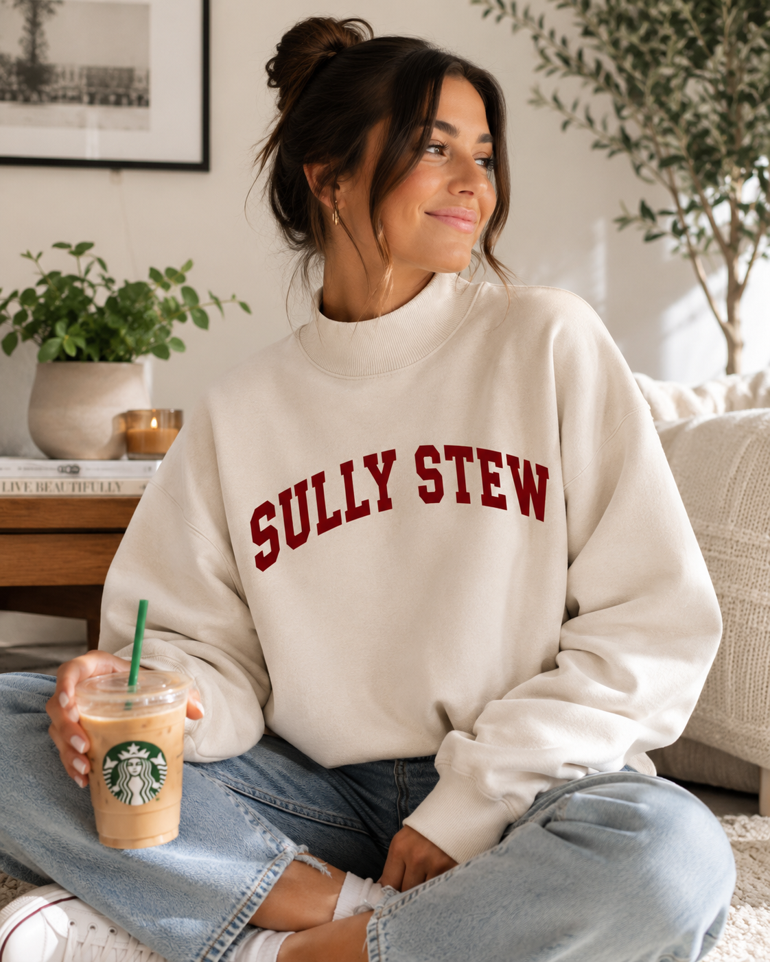A woman sitting indoors wearing a cream sweatshirt with 'SULLY STEW' printed in red across the chest, paired with light blue jeans and holding an iced coffee. The Starbucks logo is visible on the drink.
