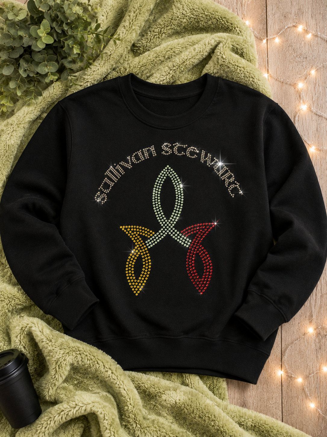 A black crewneck sweatshirt with 'Sullivan Steward' and a diamond-embellished awareness ribbon in yellow, green, and red, styled on an olive green blanket with warm fairy lights in the background.