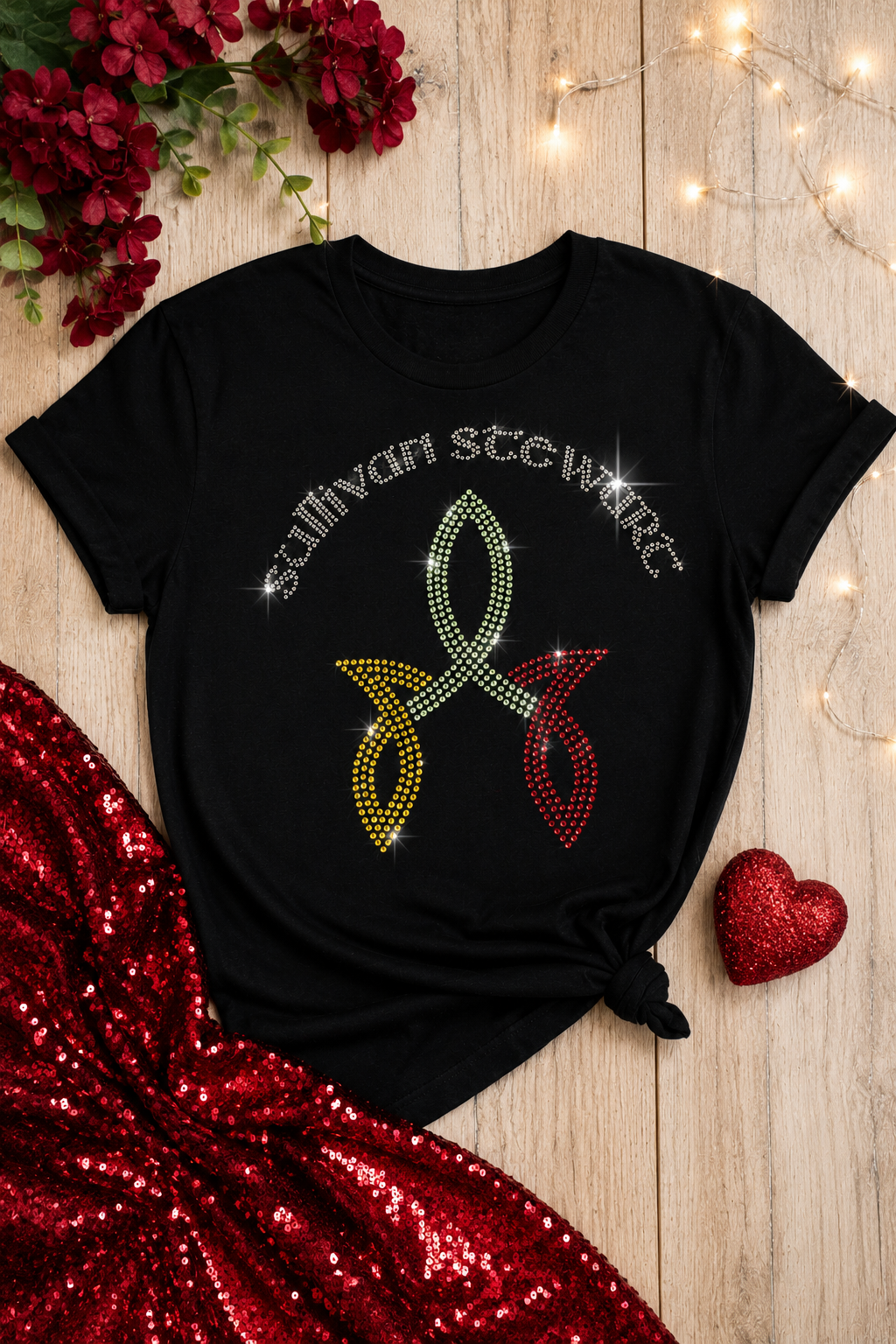A black short-sleeve T-shirt laid flat on a light wood surface, styled with red flowers, fairy lights, a red sequined fabric prop, and a red heart. The shirt has rhinestone text reading 'sullivan steward' above a ribbon design in yellow, green, and red.