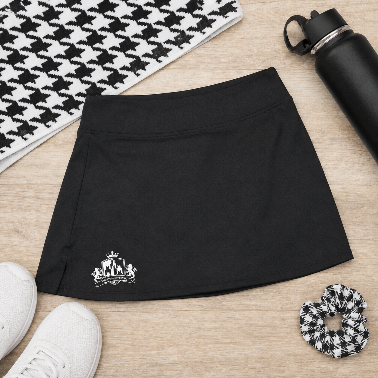 A black athletic skort with a white embroidered Australian coat of arms logo, styled in a flat lay on a light wood floor with a black and white houndstooth patterned rug, white sneakers, a black water bottle, and a black-and-white scrunchie.