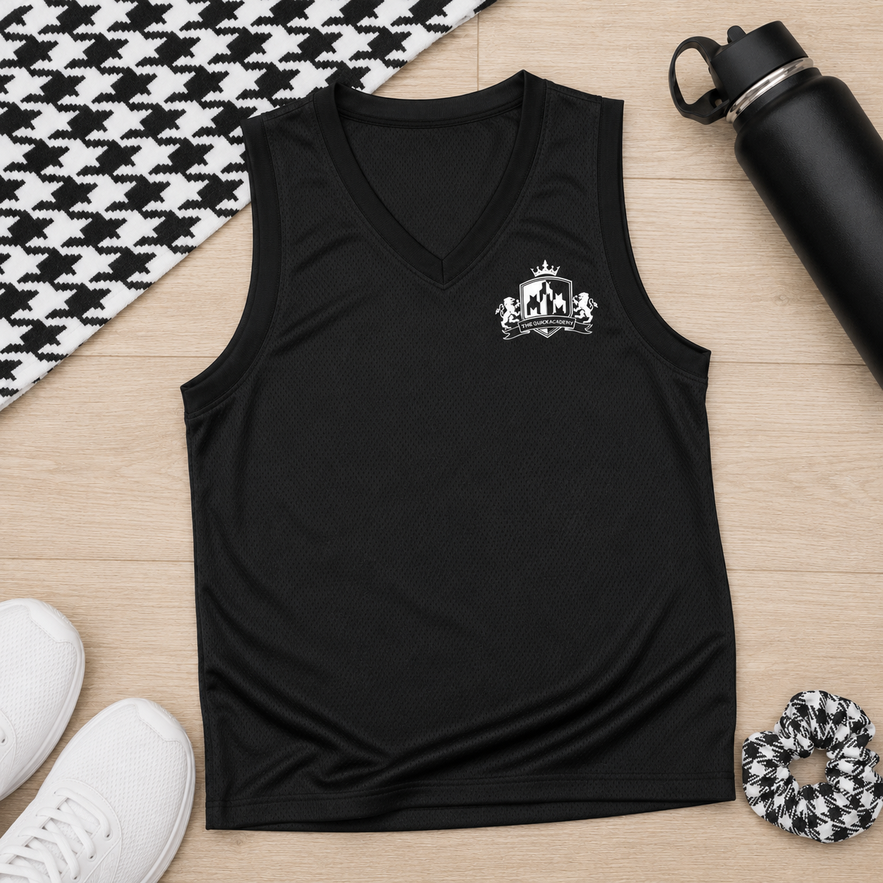 A black sleeveless V-neck athletic tank top with a small white crest graphic on the left chest, laid flat on a light wood surface with lifestyle props including a black water bottle, white sneakers, a black-and-white houndstooth fabric, and a matching scrunchie.