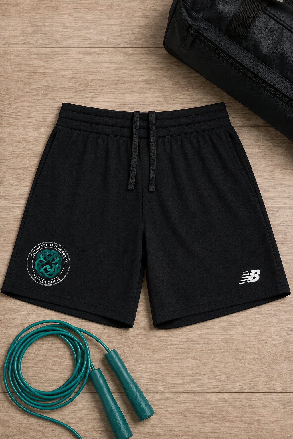 Black athletic shorts with a white New Balance logo on the left leg and a green circular Celtic logo on the right leg, laid flat on a light wood floor with a green jump rope and a black duffel bag nearby.