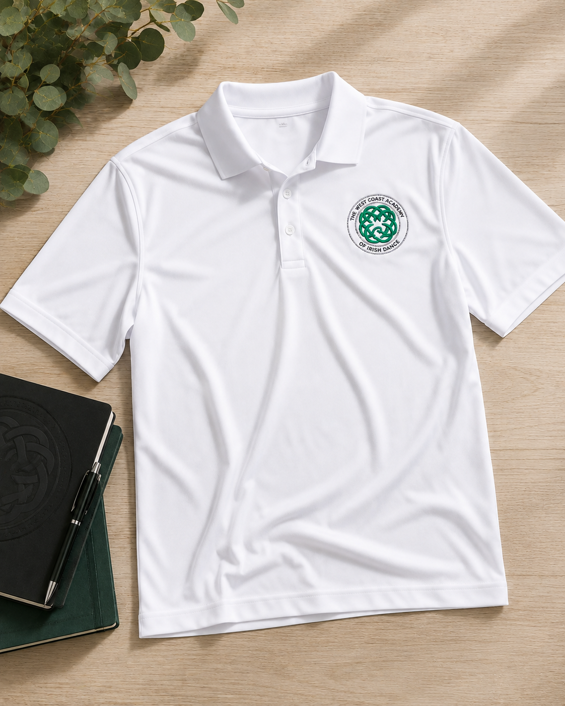 A white short-sleeve polo shirt laid flat on a light wood surface, with a green and white circular crest printed on the left chest. A black notebook and green leaves are placed nearby as styling props.