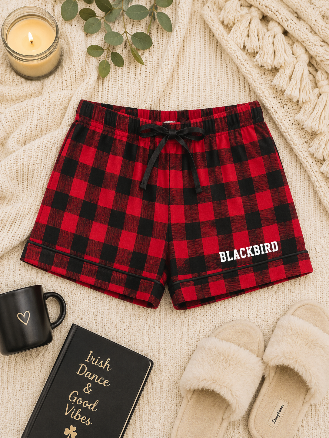 Red and black buffalo plaid pajama shorts with a white 'BLACKBIRD' text print on the left leg, styled in a cozy flat-lay arrangement with a cream knit blanket, slippers, a candle, a mug, and a notebook.