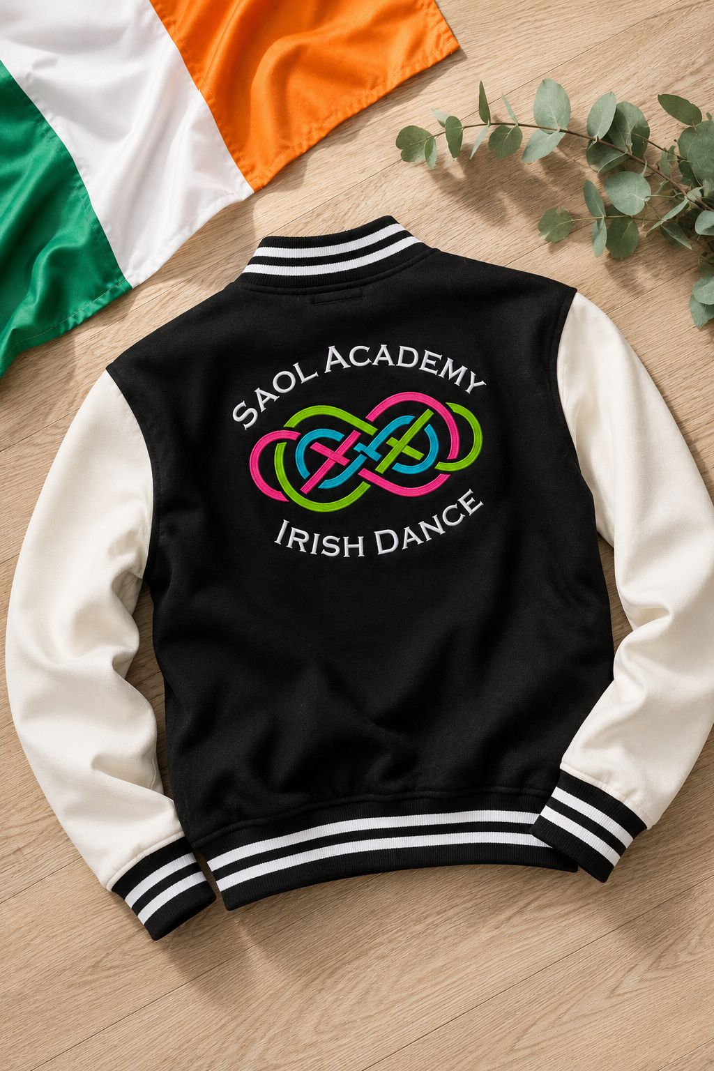 A black and white varsity jacket laid flat on a light wood surface, with a colorful 'SAOL ACADEMY IRISH DANCE' graphic on the back. An Irish flag and eucalyptus leaves are partially visible in the background.