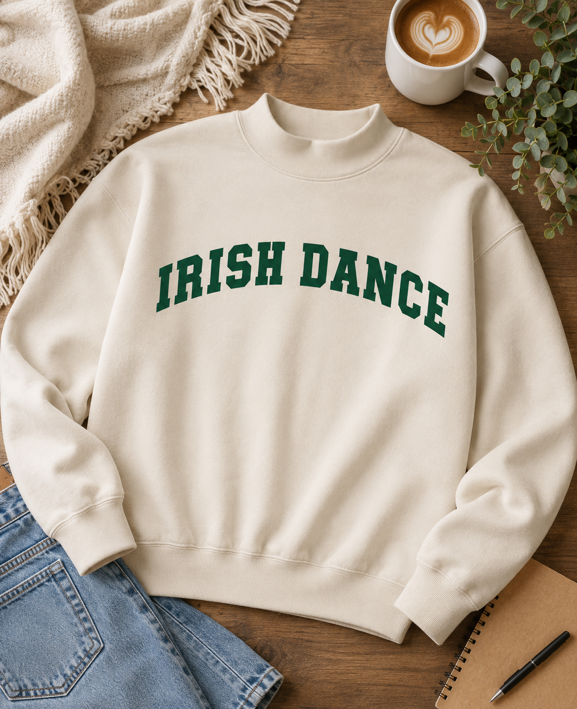 An off-white crewneck sweatshirt with 'IRISH DANCE' printed in green arched block letters across the chest, styled in a flat lay on a wooden table with a coffee cup, scarf, jeans, and notebook.