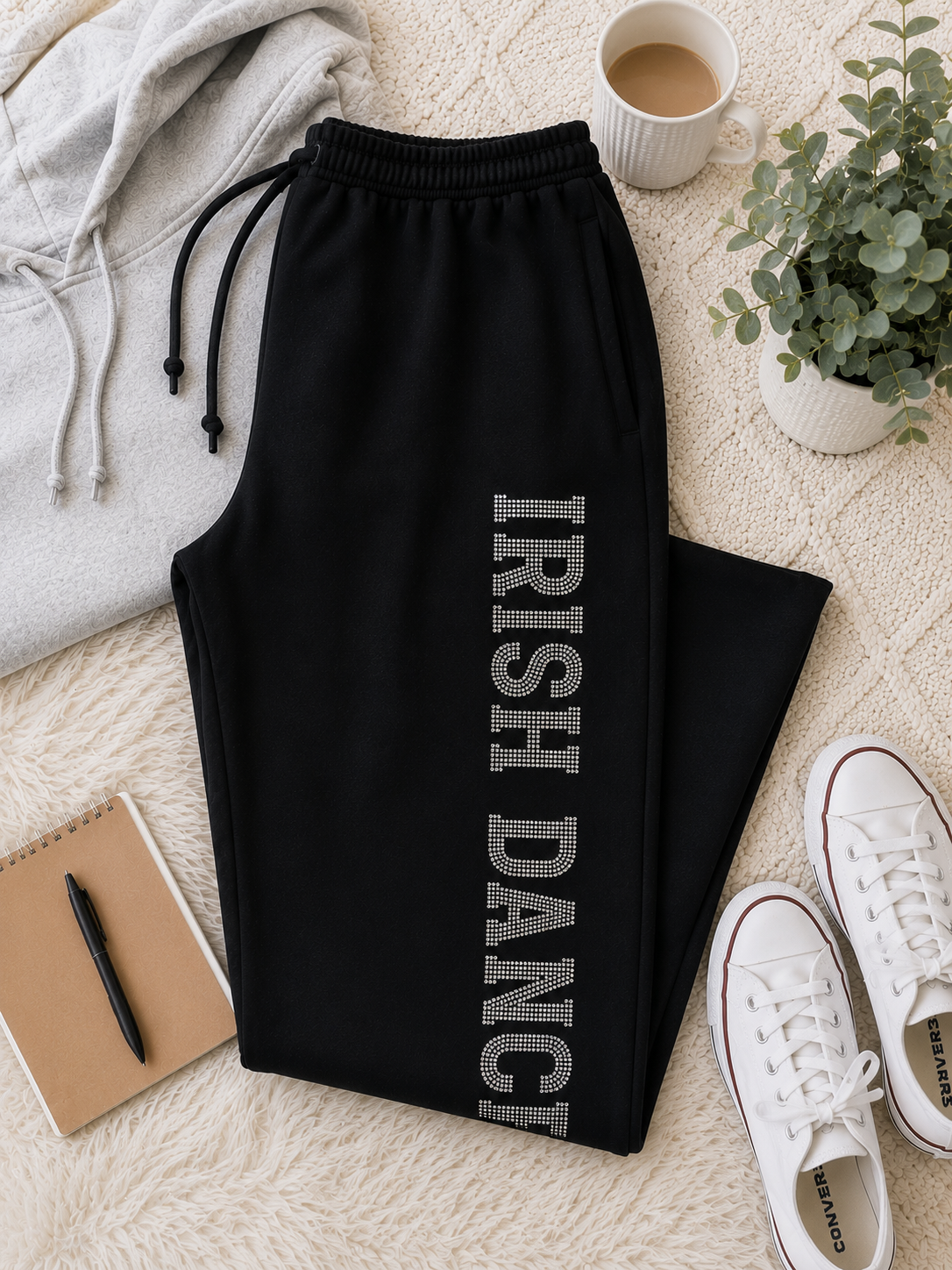 Black sweatpants laid flat on a cream textured rug, featuring 'IRISH DANCE' printed vertically in gray rhinestone-style lettering down the left leg. The pants have an elastic waistband with black drawstrings.