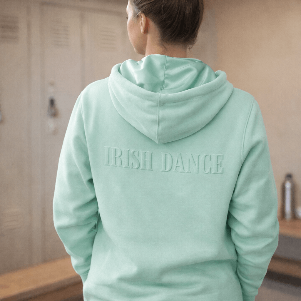 A person viewed from behind wearing a mint green hoodie with 'IRISH DANCE' embossed on the back, standing in front of wooden lockers.
