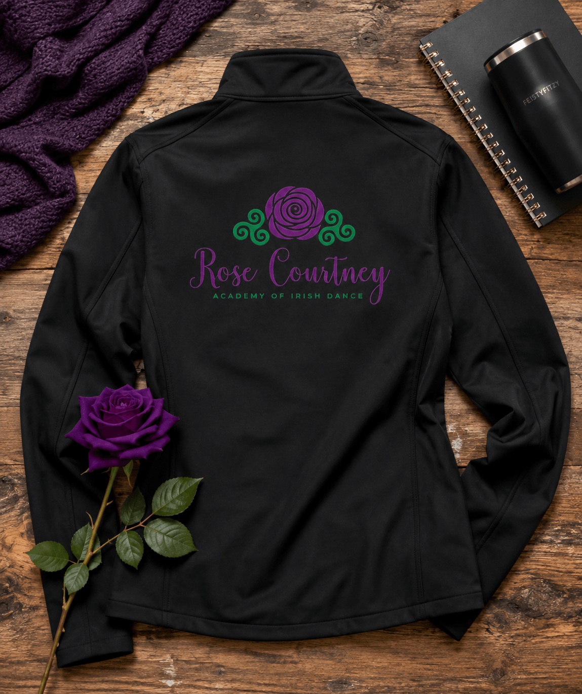 A black long-sleeve athletic jacket laid flat on a rustic wooden surface, featuring a purple rose graphic and the text 'Rose Courtney Academy of Irish Dance' printed on the back. A purple rose stem, a dark knit blanket, and a notebook with a tumbler are arranged around it as styled props.