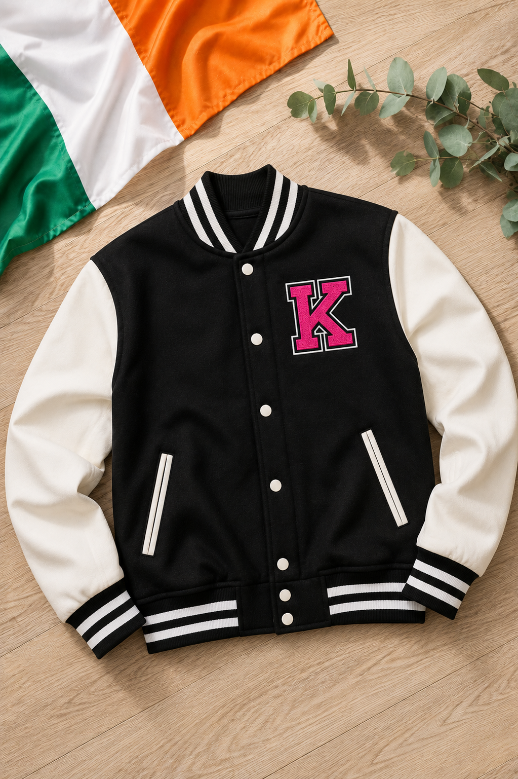 A varsity-style jacket laid flat on a light wood surface, with a green, white, and orange fabric prop in the top left and greenery in the top right. The jacket has a black body, white sleeves, white striped ribbed trim, and a large pink letter K on the chest.